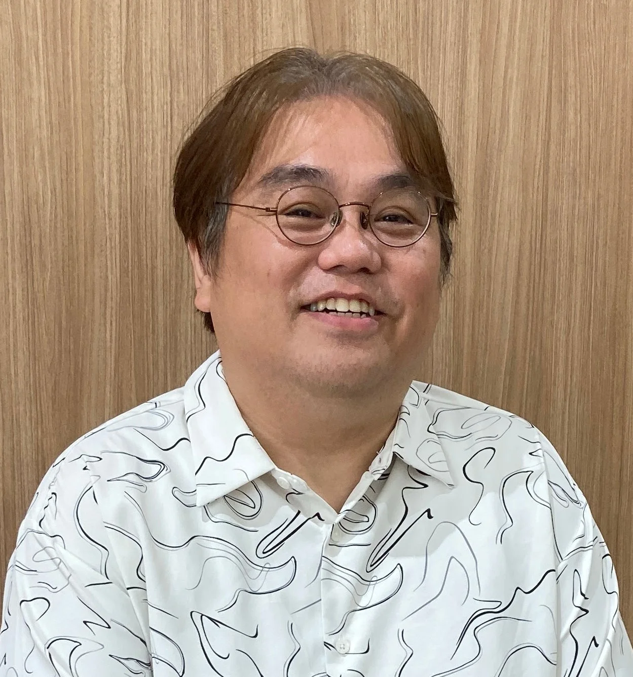 丸眼鏡をかけた笑顔のアジア人男性が、木目調の背景を背に白い柄シャツを着て写っています。