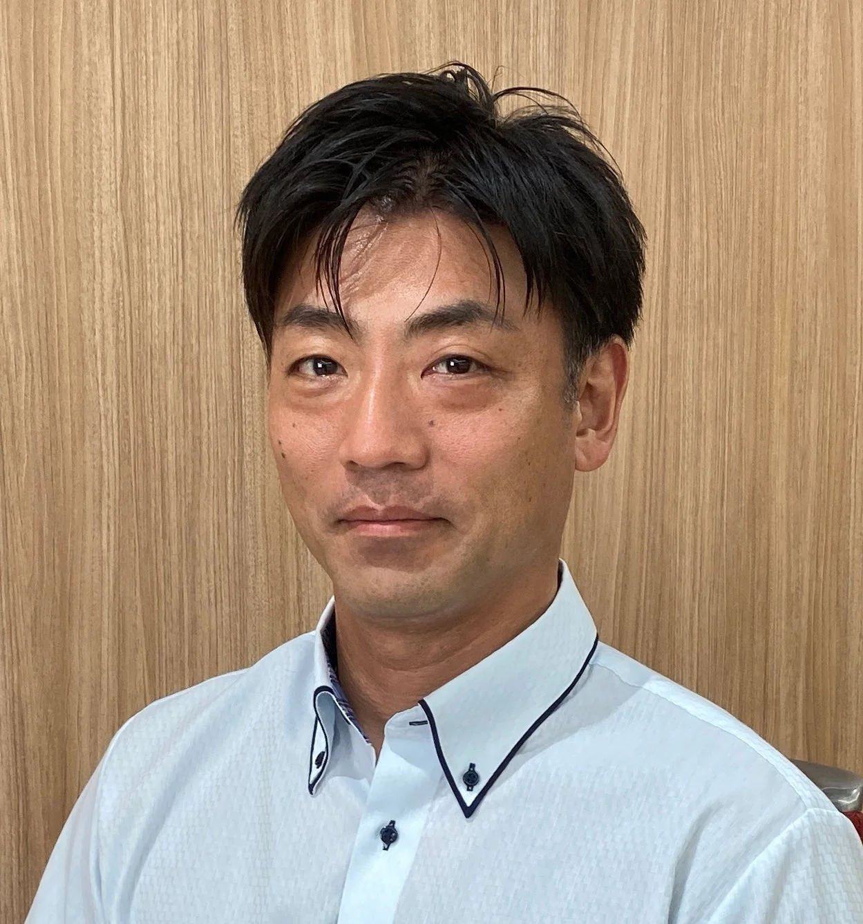 木目調の背景を背に、水色のシャツを着たアジア系の中年男性がカメラをまっすぐ見つめているポートレート写真です。