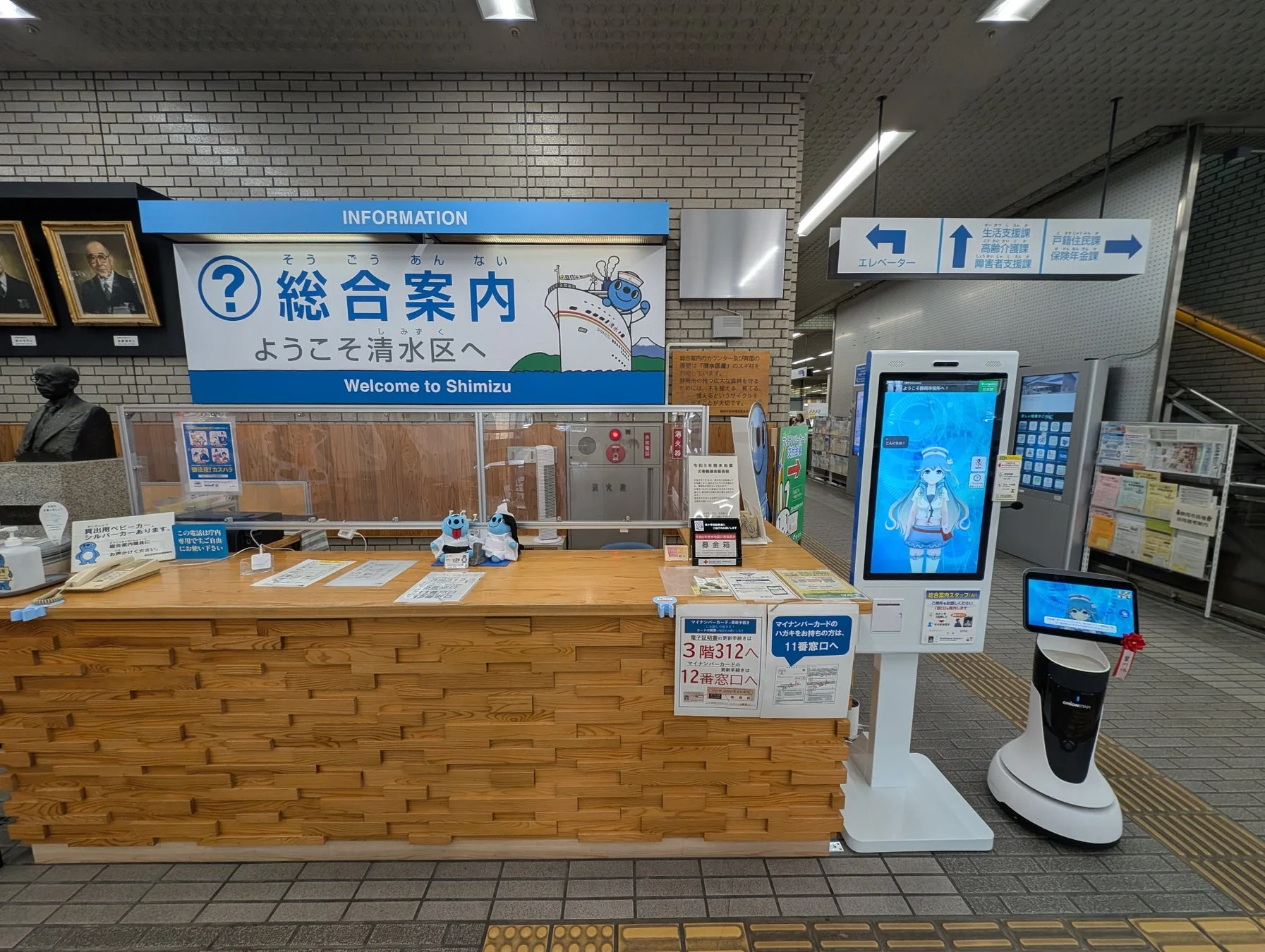 清水区の総合案内カウンターの様子。木目調のカウンターには、マイナンバーカードの手続き案内やベビーカー貸出情報がある。デジタルサイネージと案内ロボットも設置されており、効率的な情報提供がなされている。