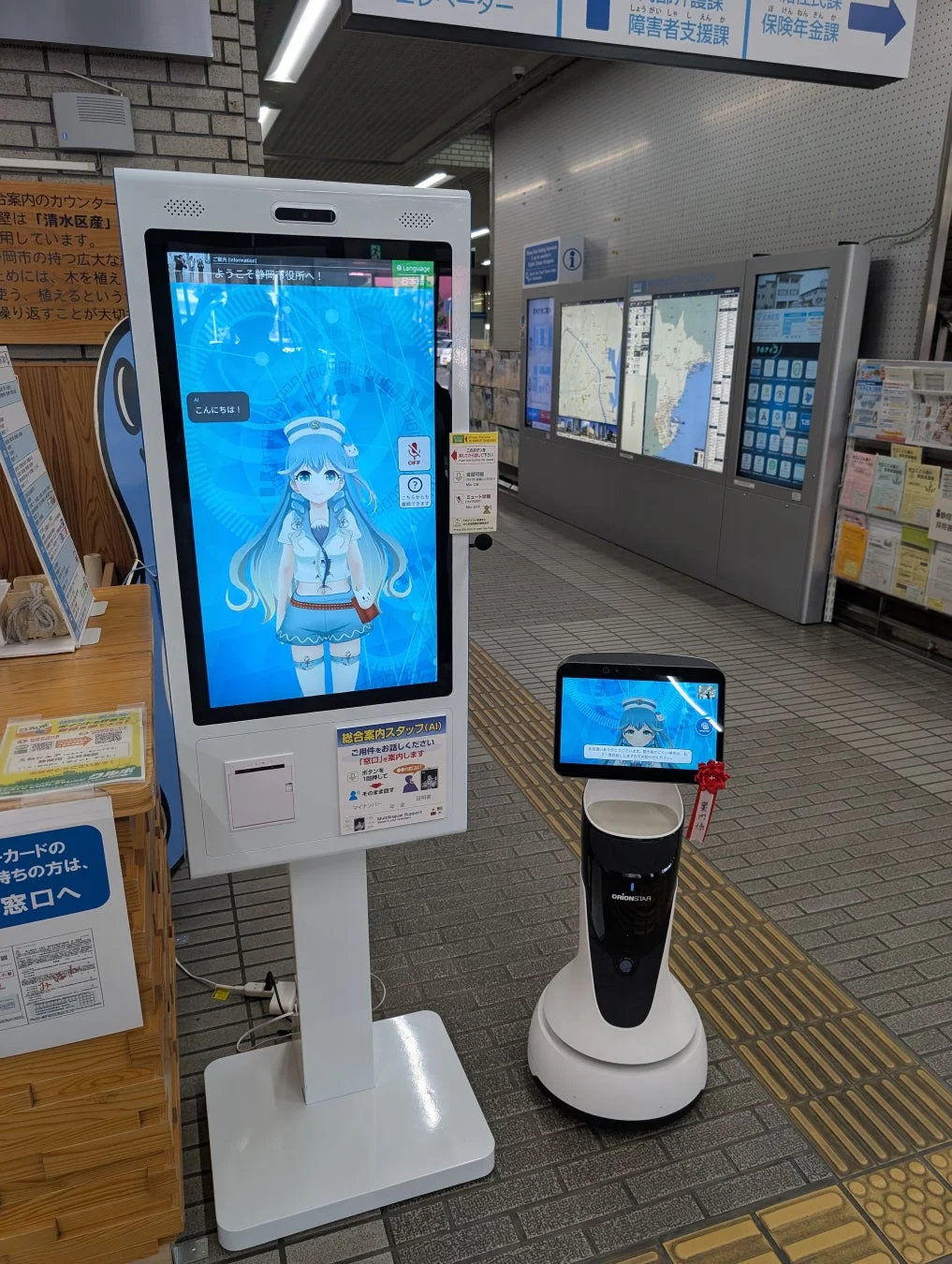 公共施設に設置された、アニメキャラクターのAIアシスタントが表示された大型デジタルサイネージと、小型のサービスロボット「OrionSTAR」が来訪者を出迎えています。現代的な情報提供システムが導入されている様子です。