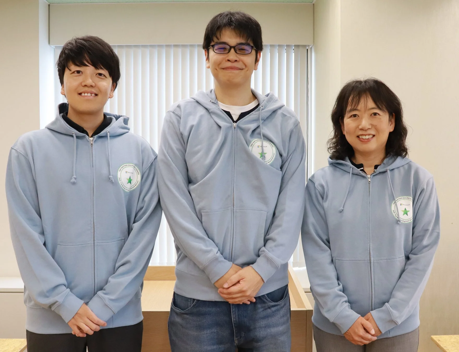 Microsoft Top Partner Engineer Awardのロゴ入り水色パーカーを着た3人が、屋内で笑顔で並んでいる集合写真